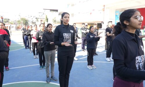 A group of young students in sports attire stand in rows on an outdoor basketball court