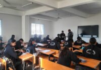 Students in a classroom wearing similar jackets, with one standing near a digital screen.